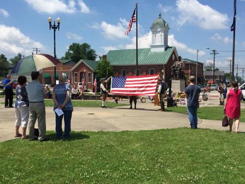 A group of people gather outdoors, holding an American flag near a statue, with a building and trees in the background.