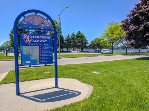Sign for Ludington's Waterfront Walkway, listing points of interest, with a clear blue sky and green grass in the background.