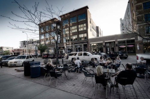 Outdoor café scene with people dining at tables, surrounded by buildings and parked cars on a sunny day.