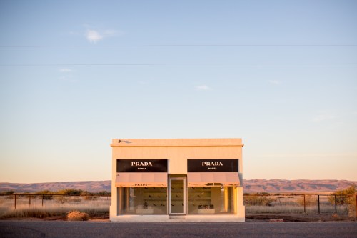 A minimalist Prada store in a remote desert landscape, featuring large windows and black awnings against a sunset sky.