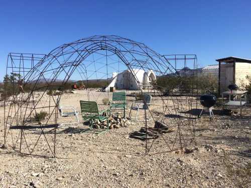 A metal frame structure in a desert landscape, surrounded by chairs and a fire pit, with distant buildings in the background.