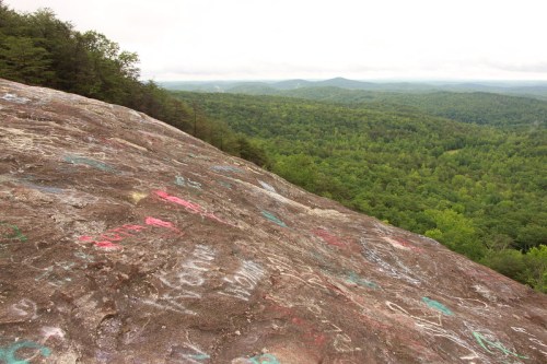 A rocky surface covered in colorful graffiti, overlooking a lush green forest and distant mountains under a cloudy sky.