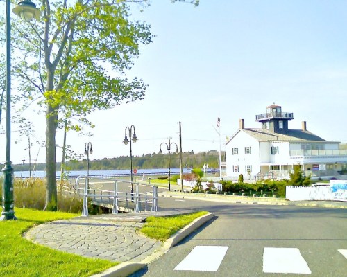 Scenic waterfront view featuring a white building, lampposts, and a pathway leading to the water. Clear blue sky.