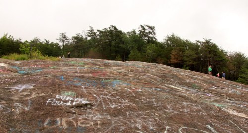 A rocky surface covered in colorful graffiti, with people exploring in the background and trees lining the area.