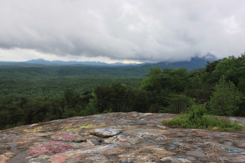 A rocky overlook with colorful graffiti, surrounded by lush green trees and mountains under a cloudy sky.