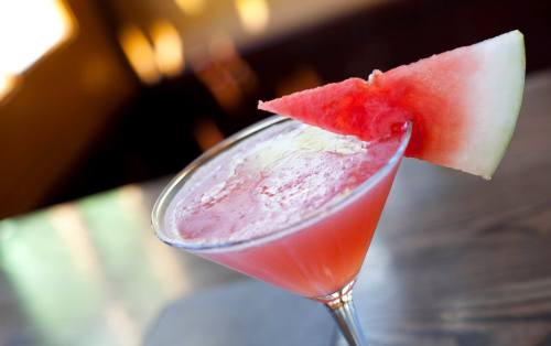 A refreshing pink cocktail garnished with a slice of watermelon, served in a martini glass on a wooden table.