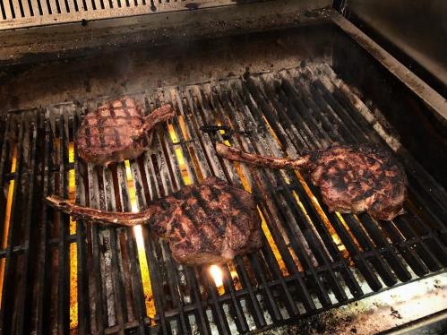 Three large, grilled tomahawk steaks sizzling on a barbecue grill with flames and grill marks visible.