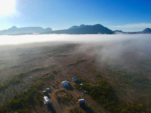 Aerial view of a campsite surrounded by fog, with mountains in the background and clear blue skies above.