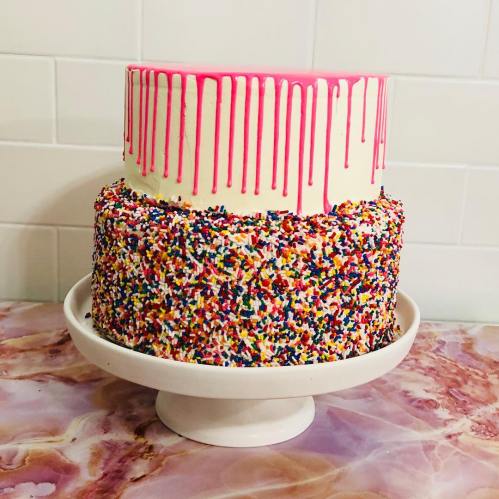 A two-tiered cake with pink drip icing and colorful sprinkles on the bottom layer, displayed on a white cake stand.