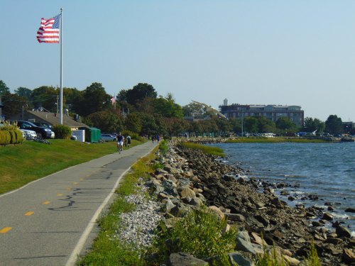 A scenic waterfront path with a flag, people walking, and a view of a building along the shore.