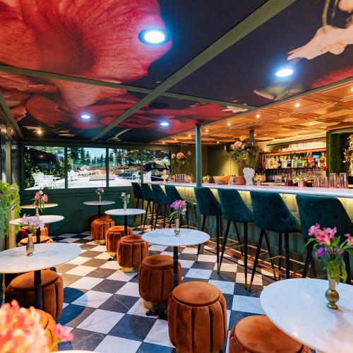 A stylish bar interior with green walls, floral ceiling, marble tables, and plush seating, featuring vibrant decor and a lively atmosphere.