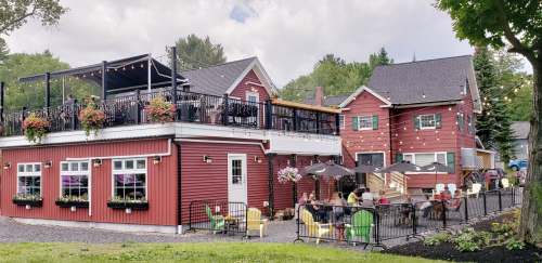 A charming red building with a rooftop deck, surrounded by greenery and outdoor seating, bustling with people.