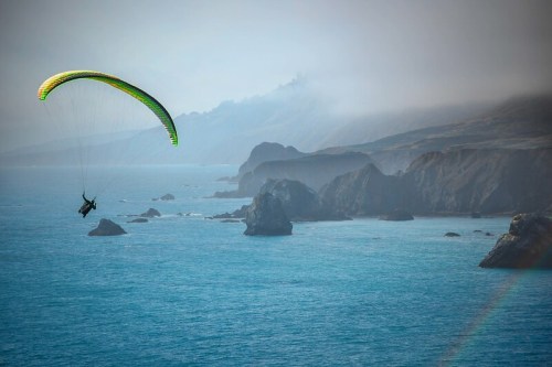A paraglider soars above a misty coastline, with rocky cliffs and a serene blue ocean below.