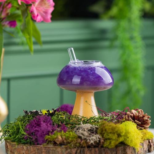 A whimsical mushroom-shaped glass filled with purple drink, surrounded by colorful moss and decorative plants.