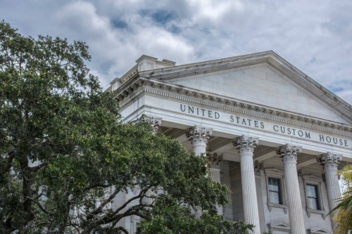 The United States Custom House, featuring classical architecture and tall columns, framed by lush greenery.