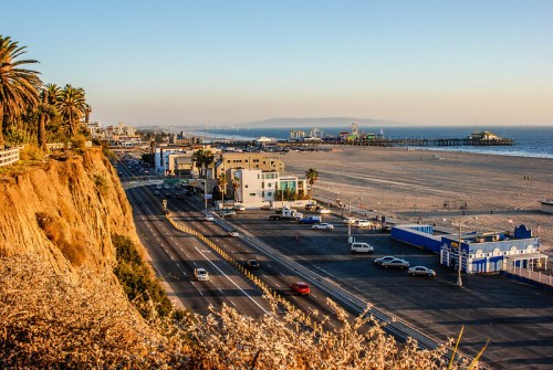 A coastal view featuring palm trees, a sandy beach, and a pier under a clear sky during sunset.