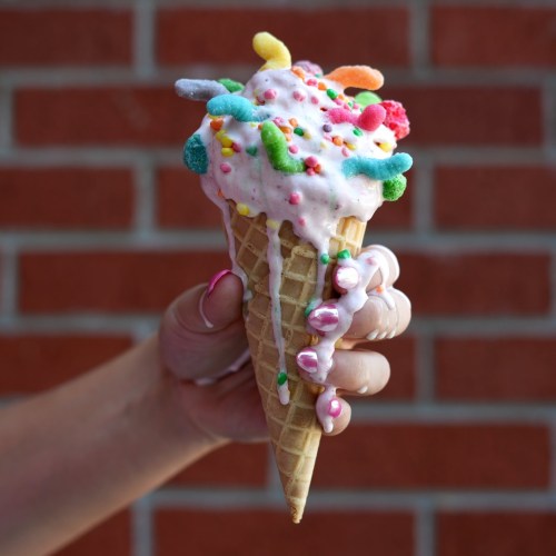 A hand holding a colorful ice cream cone topped with sprinkles and gummy candies, against a brick wall background.