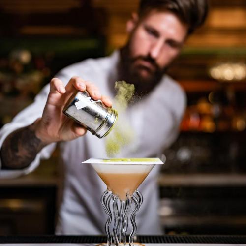 A bartender sprinkles green powder over a cocktail in a unique glass, showcasing a creative drink preparation.
