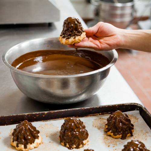 A hand holds a chocolate-dipped pastry above a bowl of melted chocolate, with more pastries on a tray in the foreground.
