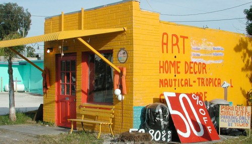 Colorful storefront with yellow walls, red door, and signs advertising art and home decor, featuring a 50% off sale.