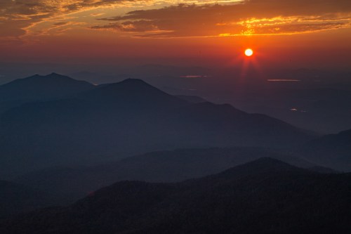 Sunset over layered mountains, with a vibrant orange sky and distant lakes reflecting the fading light.