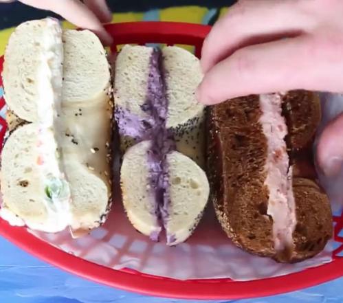 A basket with three bagels filled with different spreads: white, purple, and brown, with hands reaching for them.