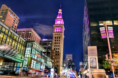 A city street at night featuring a tall clock tower illuminated in purple, surrounded by modern buildings and traffic.