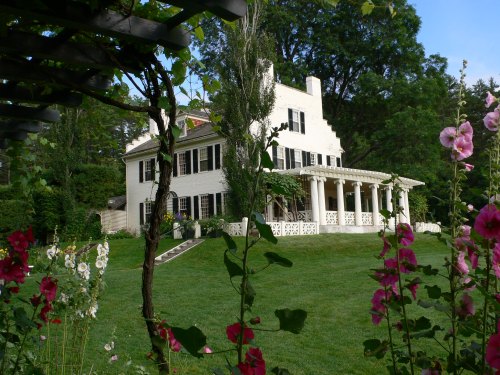 A white house with black shutters and a porch, surrounded by green grass and colorful flowers in the foreground.