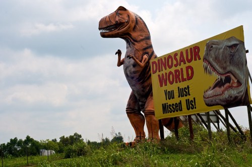 A large dinosaur statue next to a colorful sign for "Dinosaur World" with the text "You Just Missed Us!"
