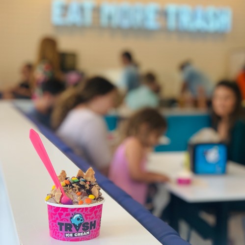 A colorful ice cream cup with toppings sits on a table, with people enjoying treats in the background.