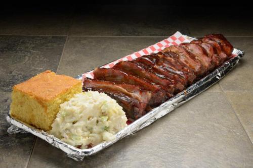 A platter featuring glazed ribs, creamy potato salad, and a slice of cornbread on a checkered paper-lined tray.