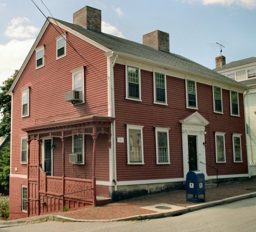 A red two-story house with white trim, featuring a porch, air conditioning unit, and a blue mailbox on the sidewalk.