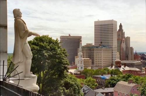 A statue overlooks a city skyline with modern buildings and historic churches under a cloudy sky.