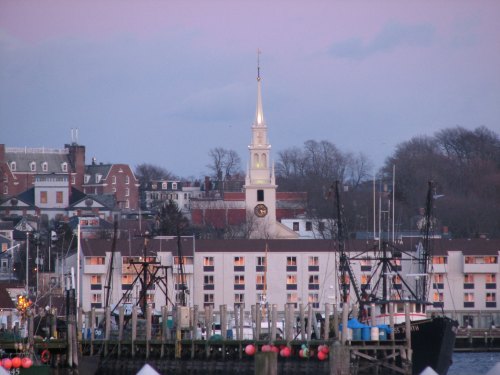 A harbor scene featuring fishing boats, a waterfront hotel, and a church steeple in the background at dusk.