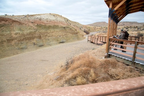 A person stands on a wooden platform overlooking a dry, rocky landscape with hills in the background under a cloudy sky.