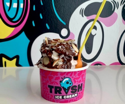 A colorful ice cream cup topped with chocolate, almonds, and a bright yellow spoon, set against a vibrant mural.