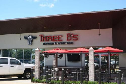 Exterior of Three B's Burger & Wine Bar, featuring a sign, outdoor seating with red umbrellas, and a white truck parked nearby.