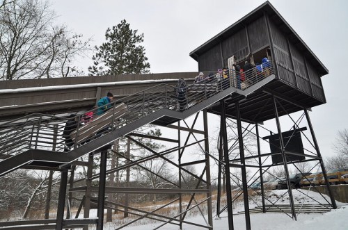 A group of people ascending a metal staircase to a wooden observation tower in a snowy landscape.