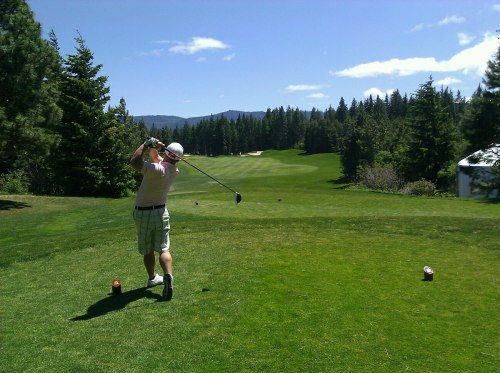 A golfer swings a club on a sunny day, surrounded by lush green trees and a scenic golf course in the background.