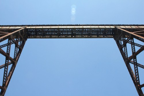 A tall, rusted bridge stretches across a clear blue sky, viewed from below.