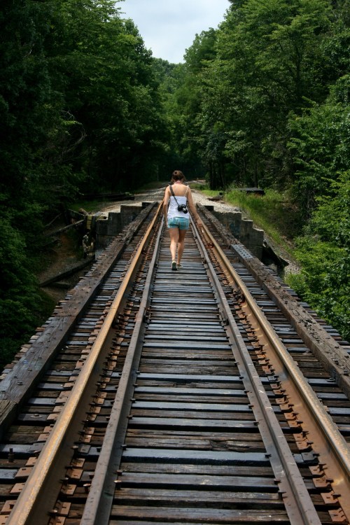 A person walks along a wooden railway track surrounded by lush green trees.