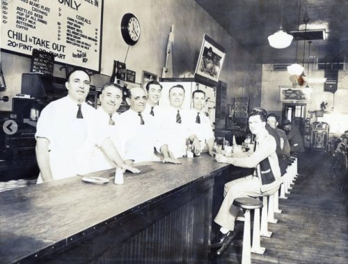 A vintage diner scene with six men in white aprons behind the counter and a seated customer at the bar.