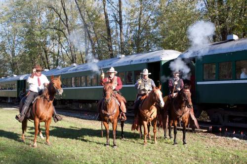 Four cowboys on horseback near a vintage train, with smoke rising from their guns in a wooded area.