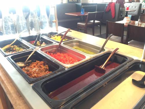 A buffet station with various toppings and sauces in metal trays, including vegetables, meats, and salsas.