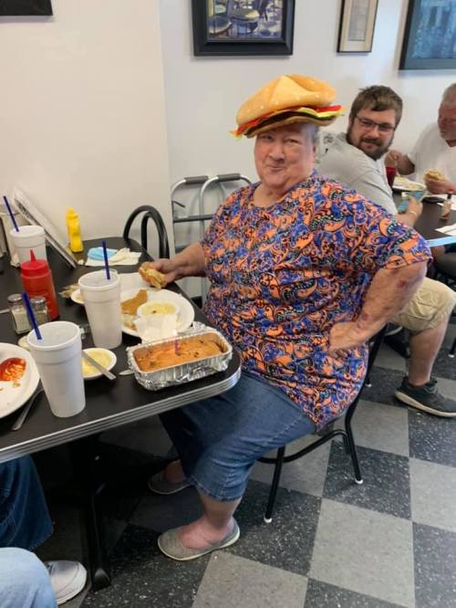 A woman wearing a burger hat smiles while sitting at a table with food in a casual dining setting.