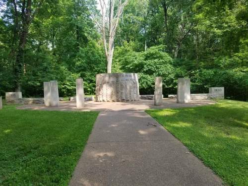 A stone monument surrounded by trees, with engraved dates and a circular structure in a grassy area.