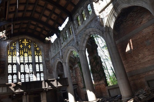 Interior of an abandoned building with tall arched windows, exposed brick walls, and overgrown vegetation.