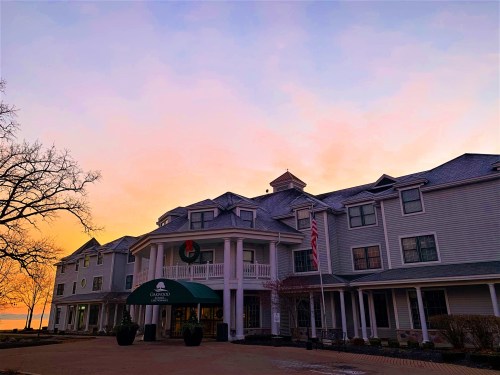 A hotel entrance at sunset, featuring a green awning and festive decorations, with trees and a lake in the background.