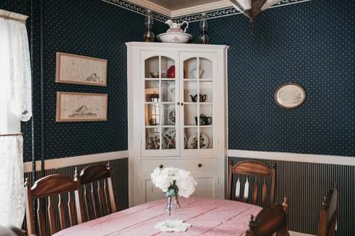 A cozy dining room with a wooden table, white china cabinet, and dark blue patterned walls. Flowers adorn the table.