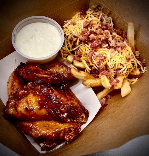A takeout box with BBQ chicken wings, loaded fries topped with cheese and bacon, and a side of ranch dressing.